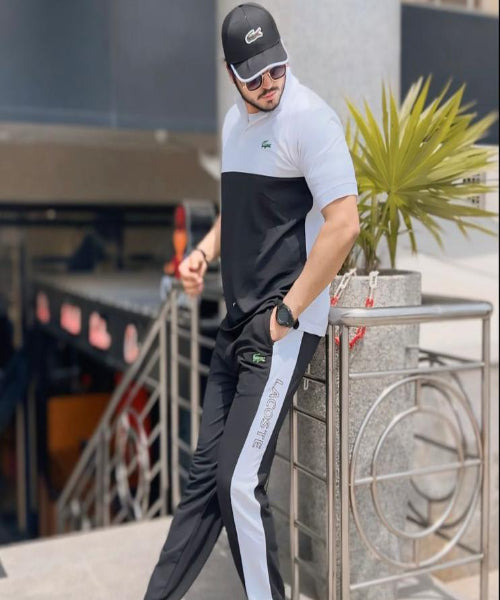 Man in black and white outfit with cap and sunglasses standing outdoors.